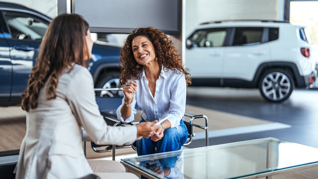 Saleswoman and a female customer in a car dealership. Sales manager explaining looking at clipboard and explaining the car features to woman customer in showroom.