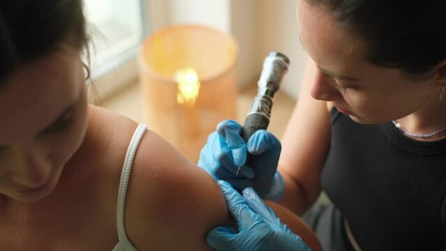 Professional tattooist makes tattoo on the shoulder. Beautiful woman in salon while tattoo artist in latex gloves working with rotor machine. Cinematic shot