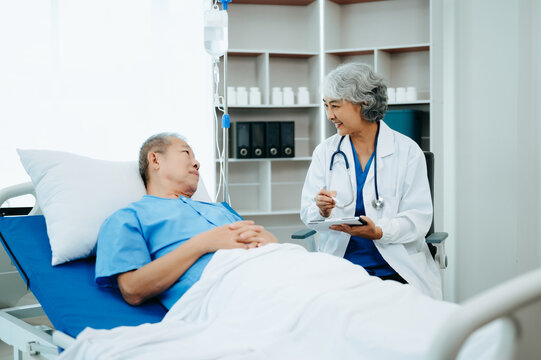 Asian Doctor In White Suit Take Notes While Discussing And Asian Elderly, Man Patient Who Lying On Bed With Receiving Saline Solution In Hospital Or Clinic.