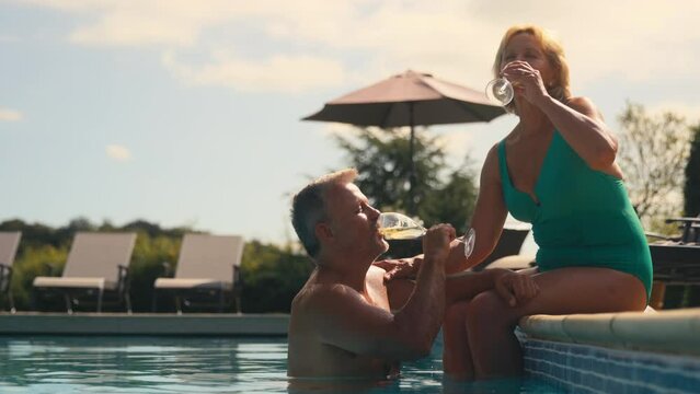 Senior retired couple on holiday with man in water and woman sitting on edge of hotel pool making toast with champagne together - shot in slow motion