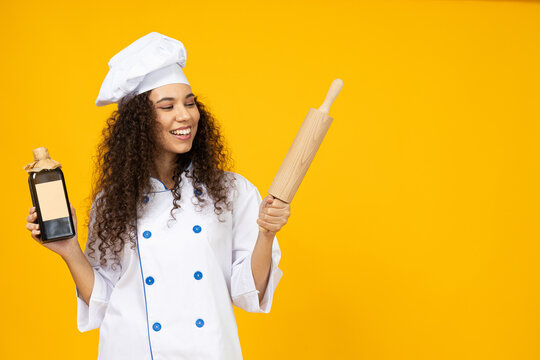 An attractive, young girl in the uniform of a chef with a rolling pin and a bottle in her hands