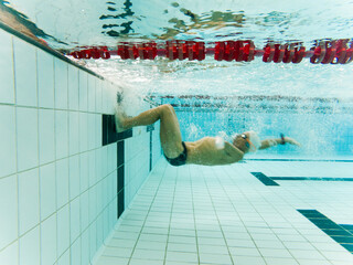 An adult man with an amputated arm is swimming underwater in a heated indoor pool wearing sports clothing. The swimmer is doing a somersault to push off the wall. Underwater photography concept.