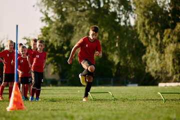 Full length of child, football player in sport uniform running, training, workout with football barriers in motion before match on soccer field.