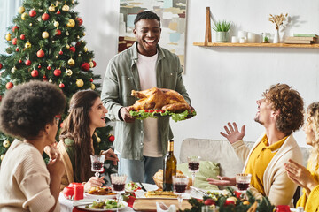joyous african american surrounded by his multicultural family holding plate with turkey, Christmas - obrazy, fototapety, plakaty