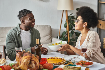 happy african american woman passing bowl with salad to relative during Thanksgiving dinner, turkey
