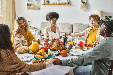joyful multicultural family members sitting and holding hands praying at festive table, Thanksgiving
