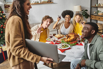 multiethnic happy family sitting at holiday lunch cheering with wine and smiling at laptop camera