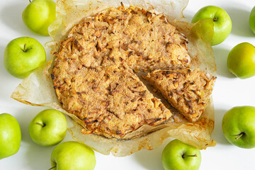 A fresh apple pie is placed on a white surface, wrapped in baking paper, and green apples lie around. A portion piece is cut off where the filling is visible.