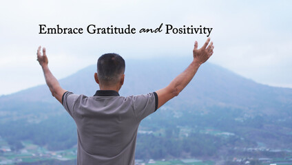 Inspirational quote - Embrace gratitude and positivity. With man standing alone from behind against the blue mountain view, raised hand and open arms. Gratefulness concept.