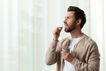 Handsome man with glass of water taking pill indoors, space for text