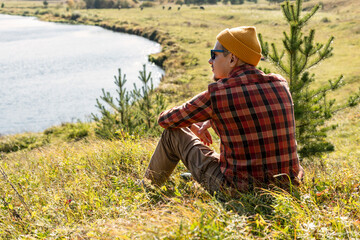 young man in red checkered shirt and beanie hat sitting on the river bank and looking at beautiful autumn view relaxing in nature tourism