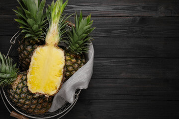 Whole and cut ripe pineapples in metal basket on black wooden table, top view. Space for text