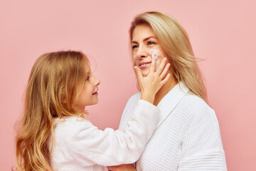 Evening hygiene. Smiling beautiful daughter, girl apply natural cosmetic cream to face, cheeks of young mother, woman over pastel pink background.