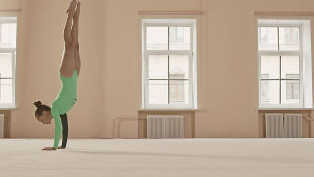 Full length side view shot of talented Caucasian gymnast girl in green leotard performing elegant floor routine with handstand, split jump and other artistic elements, rehearsing for championship