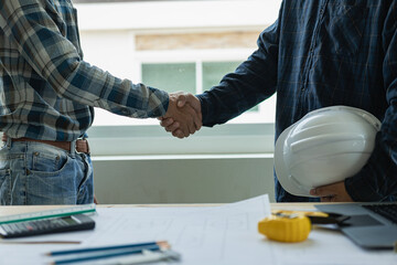 Construction workers shake hands to greet the start of a new project plan in an office center at a construction site. Partner and contractor concept with white helmets on work table
