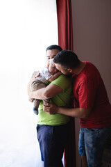 Vertical portrait of a married couple holding their adopted son near the window and gently stroking his back to relieve colic and giving him cuddles and caresses.