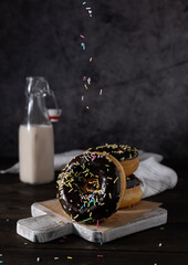 Donuts in chocolate glaze with multi-colored sprinkles on a wooden board. A bottle of melted milk in the background. Napkin on the table. Dark background