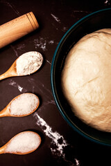yeast dough in a plate with a rolling pin and three wooden spoons with sugar, salt and flour on a black marble background