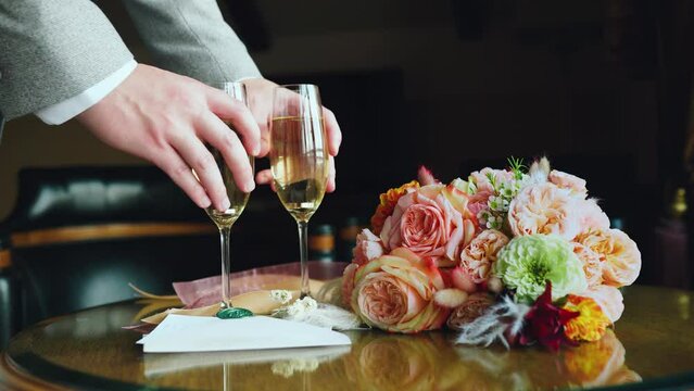 Groom takes glasses of champagne with his hand from the table where the wedding bouquet and wedding invitation lie, close-up