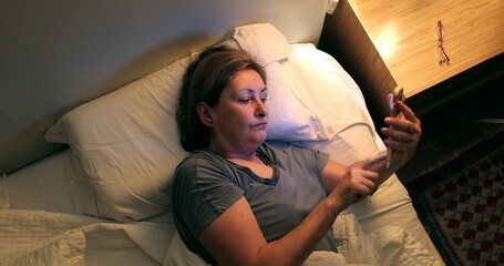 Woman picks up phone from nightstand lying in bed. Older lady browsing internet on smartphone...