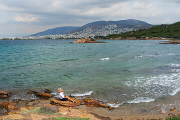 A girl sits on a stone on the Varkiza beach in Greece in autumn in windy weather.