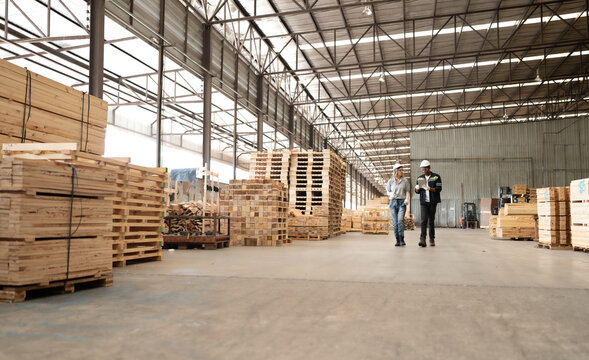 Diverse ethnic workers standing in warehouse examining hardwood material for wood furniture production. Engineer team wear safety hardhat working in lumber pallet factory. Diversity people check stock