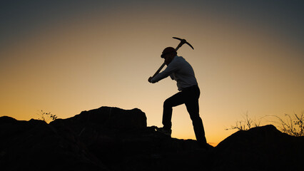Silhouette Man In Helmet With Pickaxe In His Hands Stands On Rock And Hits Stone