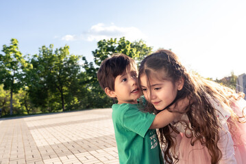 Little Brother Telling A Secret To Her Older Sister In A Park While Hugging Her At Sunset. She Is Listening Paying Attention.JPG