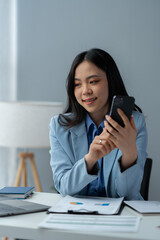 Young positive Asian female accountant using smartphone at her desk, contacting clients, checking business growth reports and relaxing on her mobile application.