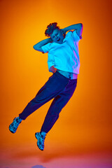 Fototapeta na wymiar Full length portrait of young attractive man jumping put his hands behind head dressed casually against gradient orange background illuminated neon light.