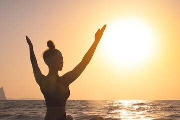 Fototapeta premium Free happy woman raises her hands and looks at the sun, rear side view. In the background, dawn, greeting the sun during yoga or meditation