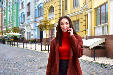 Beautiful lady in a brown coat walking on the street smiles while talking on the phone.