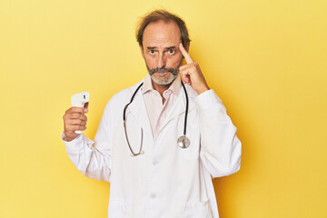 Doctor holding infrared thermometer in studio pointing temple with finger, thinking, focused on a...