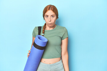 Redhead young woman holding yoga mat in studio shrugs shoulders and open eyes confused.