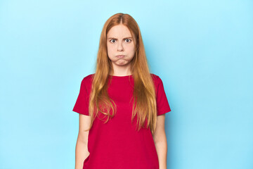 Redhead young woman on blue background blows cheeks, has tired expression. Facial expression...