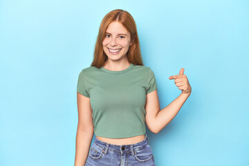 Redhead young woman on blue background person pointing by hand to a shirt copy space, proud and confident