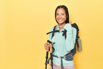 Indonesian woman with hiking gear on yellow showing a mobile phone call gesture with fingers.