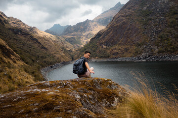 Un turista con mochila sentado con brazos abiertos en una roca a orillas de una laguna (Laguna Linda, Pachitea, Perú),paz,concepto de viajar,turismo - obrazy, fototapety, plakaty