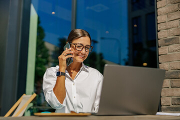 Pretty business woman talking phone while sitting on cafe terrace and working on laptop