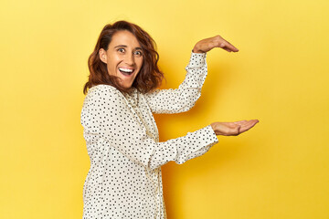 Middle-aged woman on a yellow backdrop shocked and amazed holding a copy space between hands.