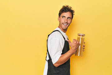 Young latino man cook with pasta jar on yellow points with thumb finger away, laughing and carefree.