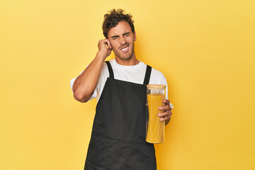 Young latino man cook with pasta jar on yellow covering ears with hands.