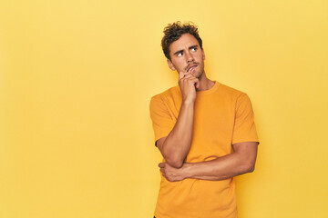 Young Latino man posing on yellow background looking sideways with doubtful and skeptical expression. - obrazy, fototapety, plakaty