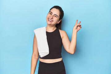 Sporty woman with towel on blue studio joyful and carefree showing a peace symbol with fingers.