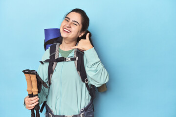 Hiker with backpack and poles on blue showing a mobile phone call gesture with fingers.