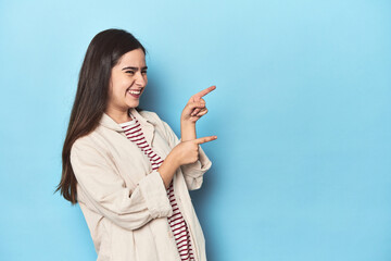 Young Caucasian woman on blue backdrop excited pointing with forefingers away.