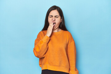 Young Caucasian woman on blue backdrop yawning showing a tired gesture covering mouth with hand.