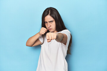 Young Caucasian woman on blue backdrop throwing a punch, anger, fighting due to an argument, boxing.