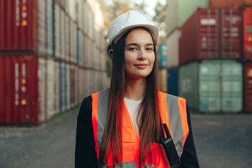 Portrait of beautiful engineer woman with walkie-talkie in white helmet and vest working in container terminal port