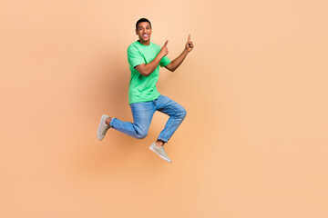 Fototapeta na wymiar Full size photo of positive guy dressed green t-shirt denim pants jumping directing at sale empty space isolated on beige color background